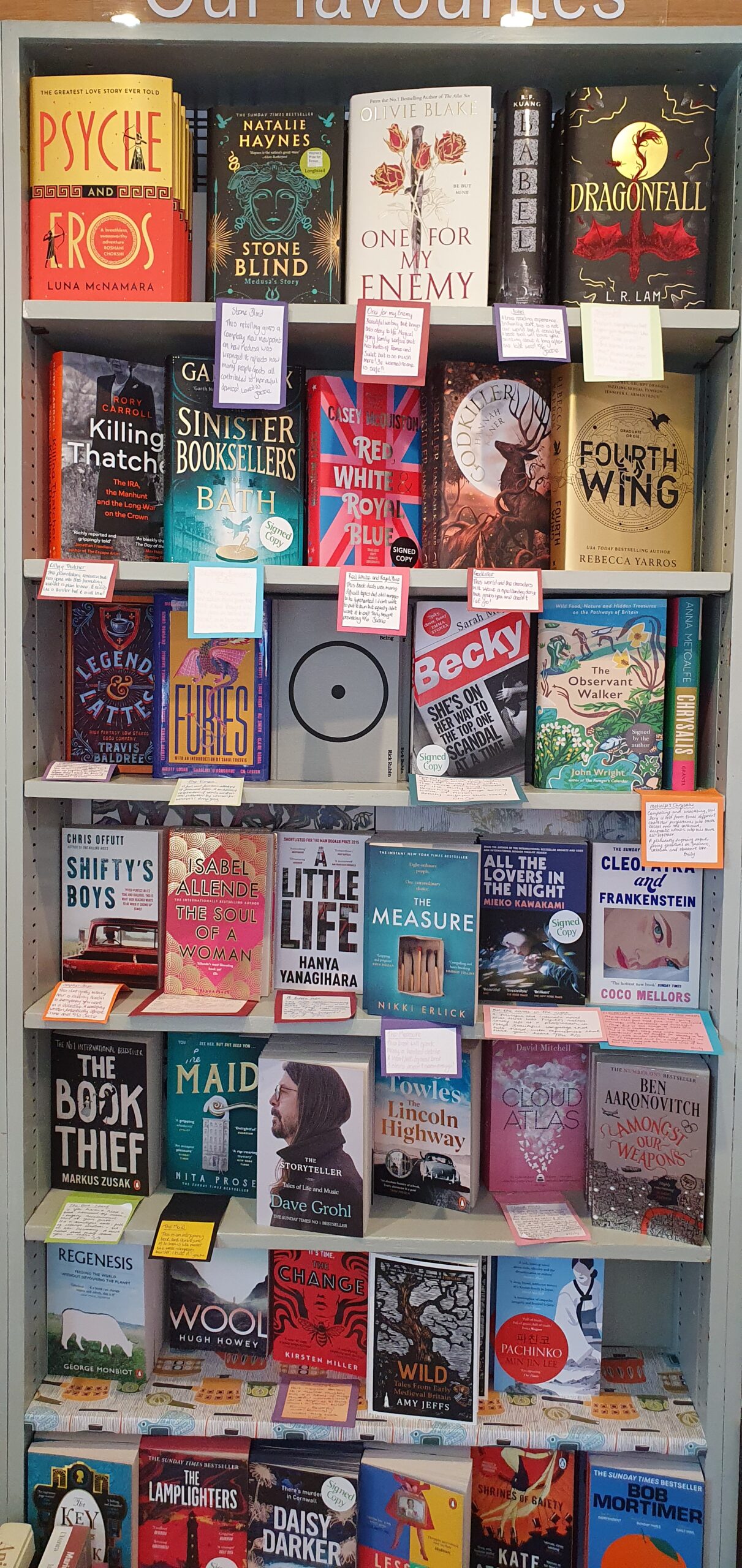An interior bookcase labelled Our Favourites, stacked four shelves high with current paperback fiction including Sinister Booksellers of Bath, Fourth Wing, Becky and Wool.