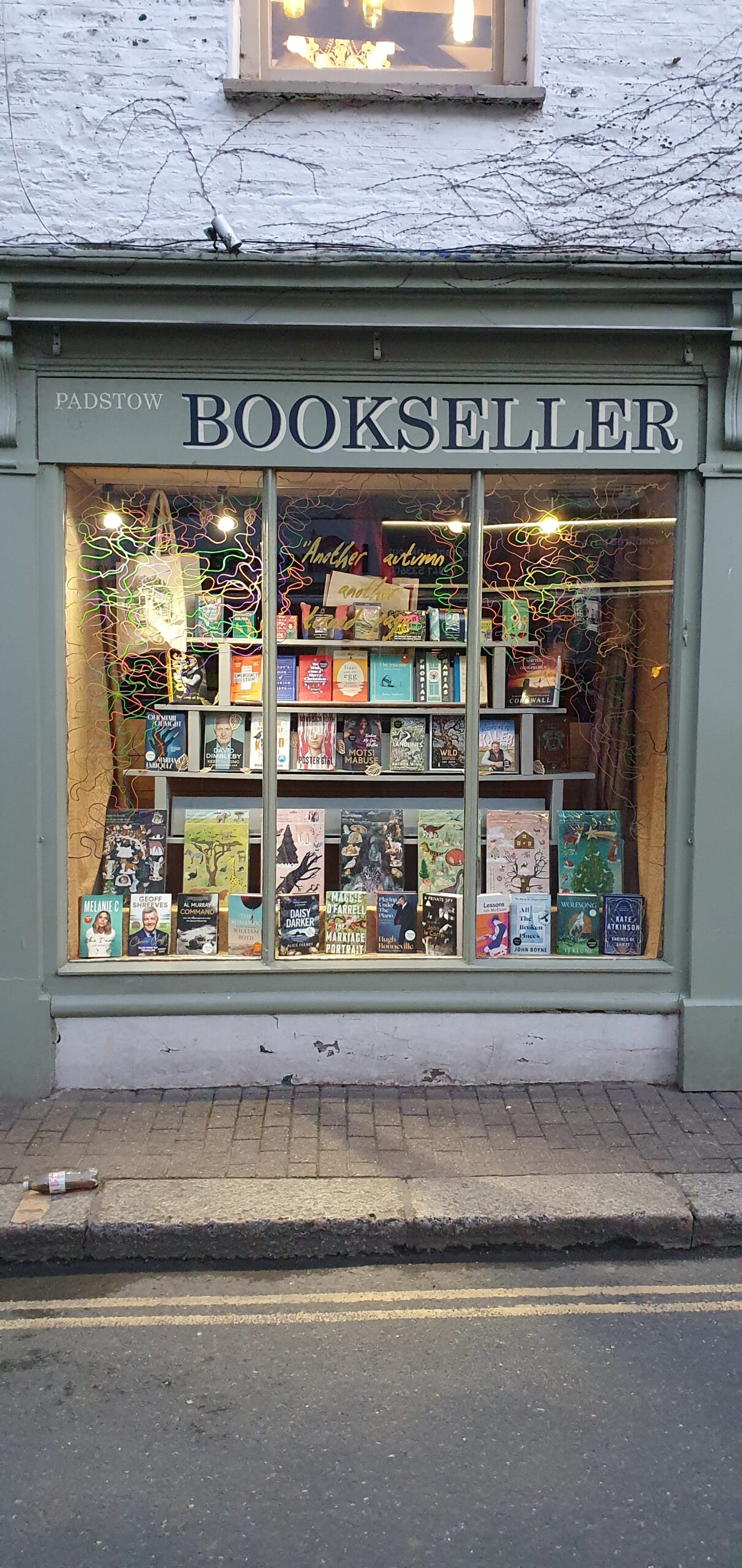 The Padstow Bookseller shopfront on Broad Street at dusk: sage-green painted woodwork with PADSTOW BOOKSELLER in serif lettering across the fascia, lit window display, books on tiered risers inside.