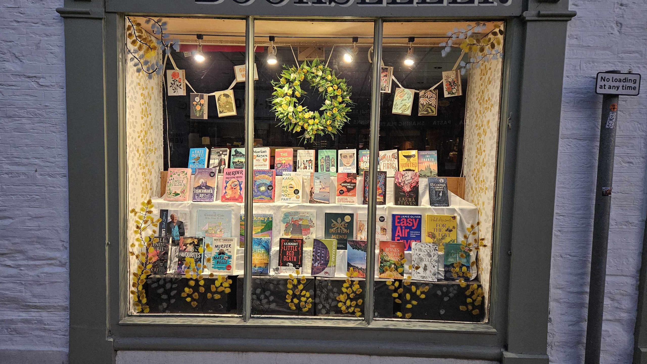 Padstow Bookseller window display at night, March: wreath of green leaves above a grid of book covers strung across the window, books arranged on a tiered riser inside.